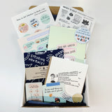 A care package titled "For anyone trying to work while the world is on fire" featuring motivational stickers, postcards, and self-care items displayed in an open box. Items include colorful sticker sheets, a bandana, art prints, and encouraging messages arranged on pastel backgrounds.