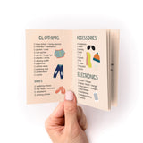 Travel Packing Checklist Zine shown open to pages displaying organized categories for clothing, accessories, and electronics. Hand holding small booklet with illustrated icons of clothes, sunglasses, and devices against peachy background. Checkbox format lists essential travel items.