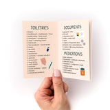 Travel Packing Checklist Zine shown open to pages displaying organized lists of toiletries, documents, and medications with cute illustrated icons, held between thumb and fingers against white background, printed on peachy-pink paper