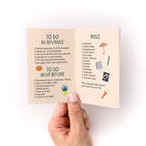 Travel Packing Checklist Zine shown open to pages with "To Do in Advance" and "Misc" sections, featuring checkboxes and cute illustrations of an umbrella, book, and other travel essentials. Printed on pink cardstock with hand-drawn style text and icons.