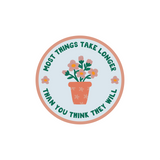 Things Take Longer Sticker featuring a circular design with a potted flowering plant in coral and pastel colors. Text curves around the edge reading Most Things Take Longer Than You Think They Will. Decorative floral illustration on pale blue background with coral border.