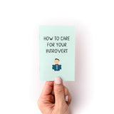 How to Care For Your Introvert Zine shown on mint green cardstock, featuring a minimalist illustration of a person reading a book. Hand with neutral manicure holds the small zine against white background, demonstrating its pocket-sized dimensions.
