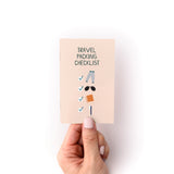 Travel Packing Checklist Zine shown as a small peach-colored booklet held against a white background, featuring a simple illustration of essential travel items including pants, sunglasses, notebook, and pen with checkmarks beside them.