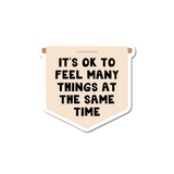 Its OK To Feel Many Things Banner Sticker featuring bold black text on a cream-colored pennant-shaped vinyl decal with rounded corners. The motivational message appears in chunky block letters against a soft background, perfect for personalizing laptops and water bottles.