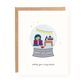 Holiday card featuring a cozy snowglobe scene with a person bundled in a blanket, reading a book. The scene includes festive string lights, a stack of books, and a warm mug, capturing a cozy holiday atmosphere.