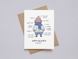 Holiday card showing a hand-drawn illustration of a person bundled in multiple layers of winter clothing including a beanie, scarf, mittens, sweaters, and boots, with labels pointing to each item and the message "Happy Holidays! Stay Cozy" on a white background, accompanied by a brown kraft paper envelope.