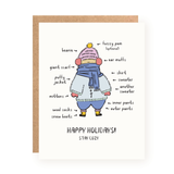 Holiday greeting card featuring a whimsical illustration of a person bundled up in multiple winter layers, labeled with various cozy clothing items like a beanie, scarf, mittens, sweaters, and boots, with the message "Happy Holidays! Stay Cozy" on a white background and accompanied by a kraft paper envelope.