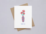 White thank you card displaying a tall blue and pink checkered vase holding four red flowers with green leaves, paired with a brown kraft envelope on a light background.