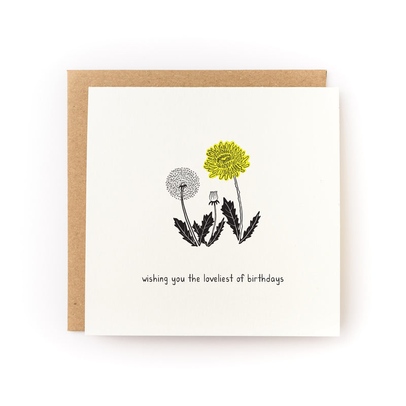 Loveliest Birthday Dandelion Letterpress Card featuring a minimalist design with two dandelions - one illustrated in black and white, one in yellow - with detailed stems and leaves on cream paper, paired with a kraft envelope. Hand-lettered text reads "wishing you the loveliest of birthdays