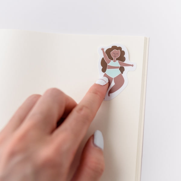 Photo of a hand holding a cute vinyl sticker.  The sticker has an illustration of  a woman dancing.