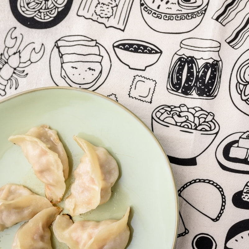 Close-up view of Home is Where the Food is Tea Towel with a plate of dumplings in the corner.