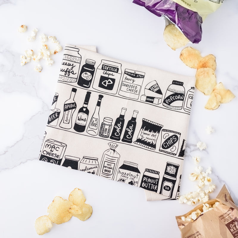 Folded up Pantry "Essentials" tea towel with funny illustrations of wine, spritzers, tomato sauce, and more -- surrounded by lots of yummy snacks.