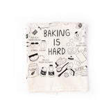 Folded up "Baking is Hard" tea towel with silly illustrations around it about how to bake, featuring different baked goods and ingredients.