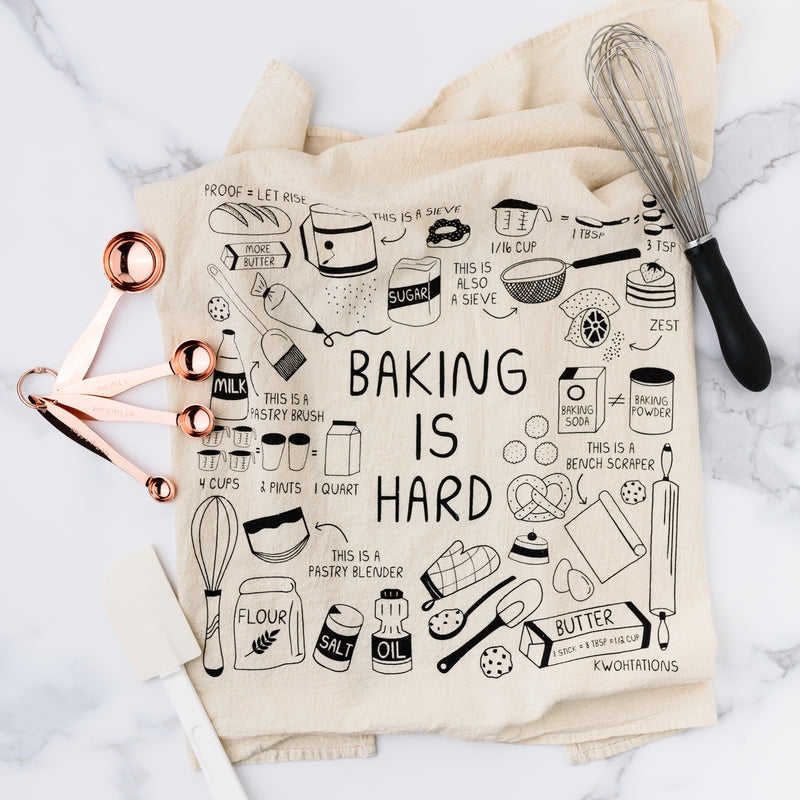 Cotton Tea Towel with "Baking is Hard" written across it with different cooking supplies around the towel