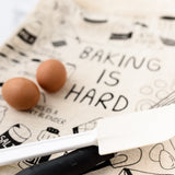 Blurred picture of "Baking is Hard" tea towel featuring a spatula, a whisk, and a couple brown eggs.