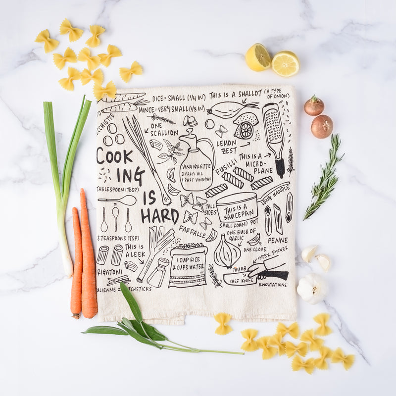 Cooking is Hard tea towel with cute drawings of lemon zest, what a saucepan is, penne pasta, and more surrounded by vegetables, pasta, herbs, and fruit.