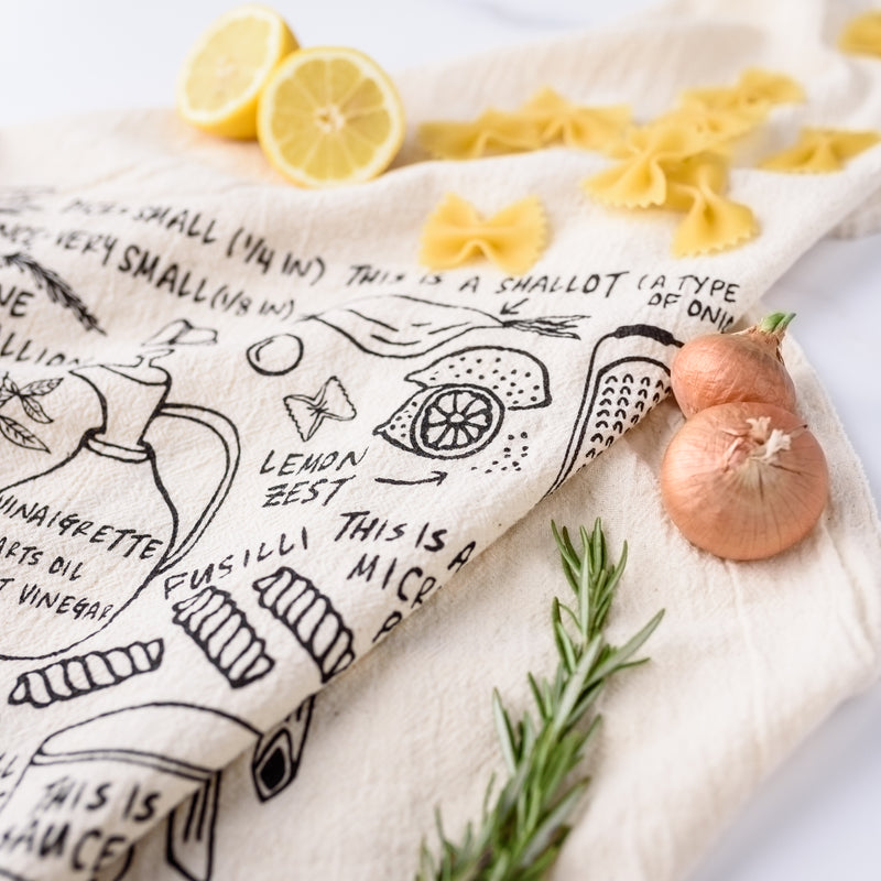 Close up view of Cooking is Hard tea towel with different pastas, how to make vinaigrette, and a shallot. Surrounded by herbs, vegetables, fruits, and pasta