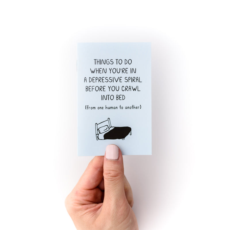 Hand holding small blue booklet about mental health titled "Things to do when you're in a depressive spiral before you crawl into bed (from one human to another)"