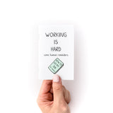 Hand holding small white booklet, which is a fun self help guide on self care at work, titled "Working is hard"